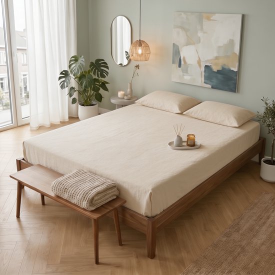 Un drap beige sur un lit en bois avec une couverture tricotée dessus. À côté, une plante et un miroir mural.