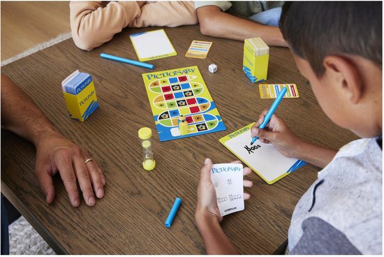 Familie speelt Pictionary spel aan tafel met spelbord, kaarten, zandloper en tekenbord.