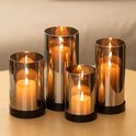 Vier rookgrijze glazen kaarshouders met zwarte bases, elk met een brandende oranje kaars, op een houten tafel.