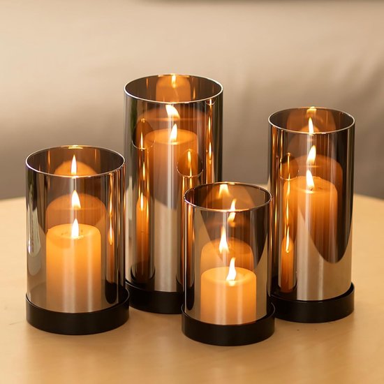 Vier rookgrijze glazen kaarshouders met zwarte bases, elk met een brandende oranje kaars, op een houten tafel.