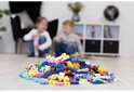 Een hoop kleurrijke Plus-Plus bouwstenen op de vloer voor een scherpe focus, met twee kinderen op de achtergrond.
