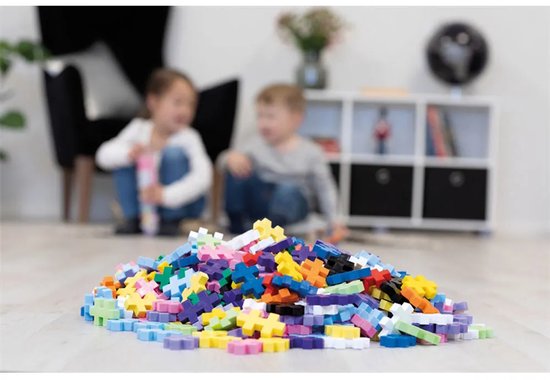 Een hoop kleurrijke Plus-Plus bouwstenen op de vloer voor een scherpe focus, met twee kinderen op de achtergrond.