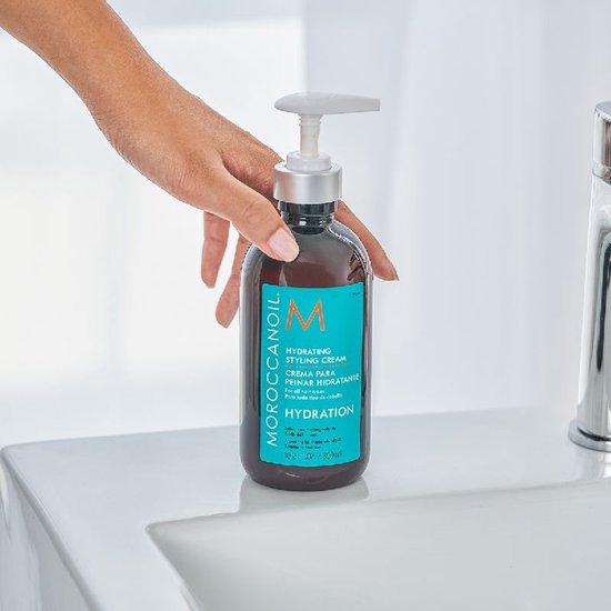 Hand drukt pompdispenser van Moroccanoil Hydrating Styling Cream fles. Fles staat op een wastafel.