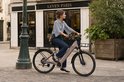 Vrouw met spijkerbroek op een champagne gouden elektrische fiets met mand voorop en leren tasje.