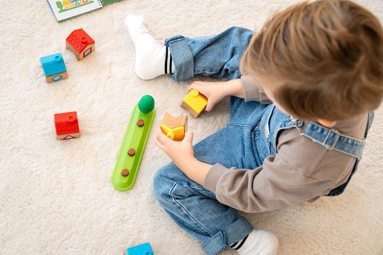 Een jongetje speelt met houten huisjes en blokken op een wit tapijt. Losse rode en blauwe huisjes, een gele blok en een groene staaf.