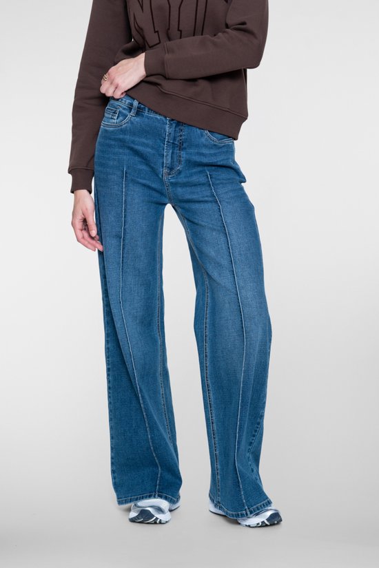 Gedetailleerde close-up van een blauwe jeans met wijde pijpen en opvallende deelnaden, gedragen door een model met een donkerbruine sweater.