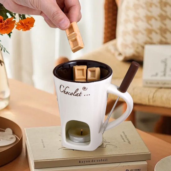 Witte mok met 'Chocolat...' tekst, gevuld met stukjes chocolade en een waxinelichtje. Een hand dip een stukje chocolade in de mok.