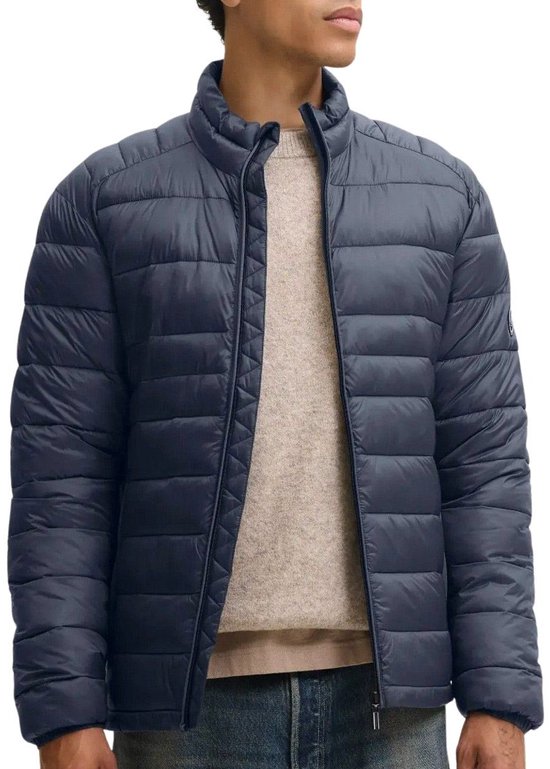 Model in een donkerblauwe, doorgestikte pufferjas met opstaande kraag, beige trui en spijkerbroek.