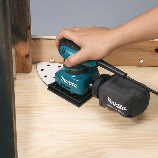 Hand die een turquoise en zwarte Makita schuurmachine vasthoudt op een houten ondergrond.
