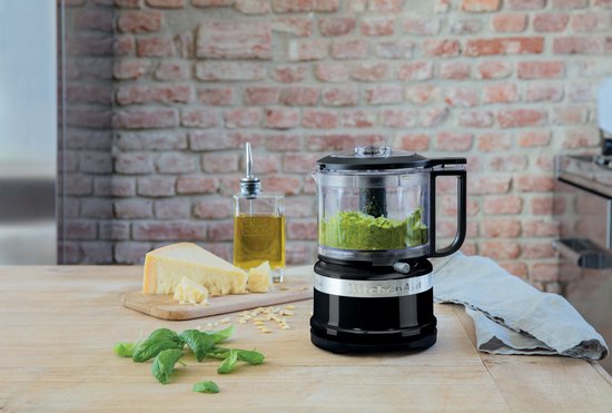 Zwarte KitchenAid hakmolen met pesto in de kan, naast basilicum en Parmezaanse kaas.