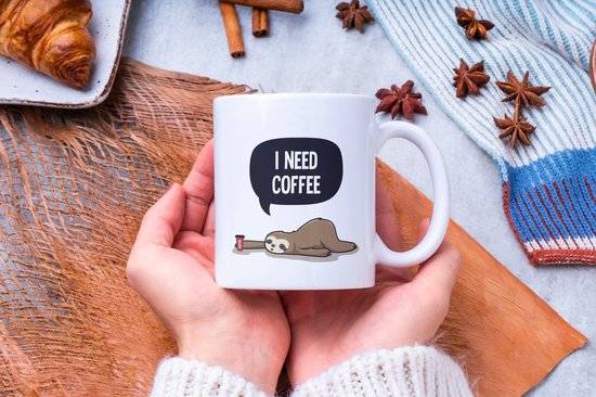 Tasse blanche avec texte 'I NEED COFFEE' et paresseux, tenue par des mains.