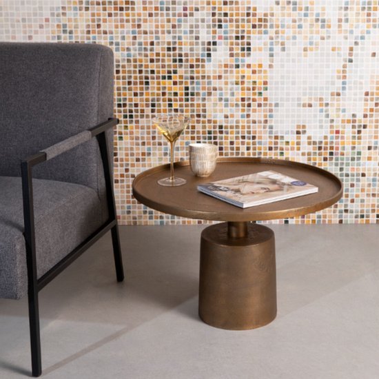 Bronzen salontafel met een ronde bovenkant, naast een grijze fauteuil, met een glas en tijdschrift.
