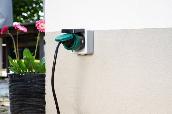 Slimme stekker in gebruik buiten, aangesloten op een muur. Naast de stekker staat een plantenbak met bloemen.