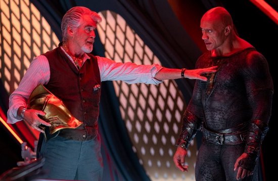Pierce Brosnan met grijze baard en vest, houdt een gouden helm vast en praat met Dwayne Johnson in een zwart superheldenpak.
