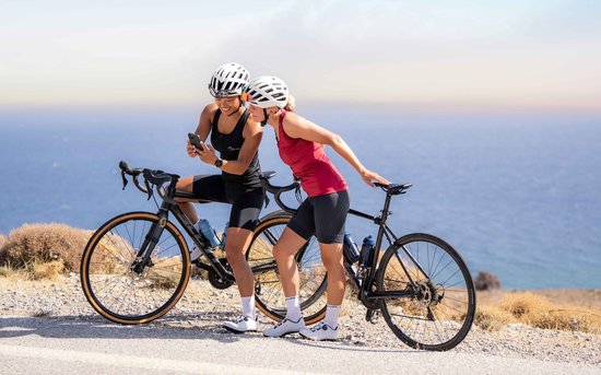 Twee vrouwen met racefietsen kijken naar een smartphone. De linker vrouw draagt een zwarte fietstop, de rechter een rode.