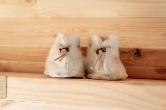 Een paar witte, wollen babyslofjes met bruine veterdetails en metalen knoopjes, staand op een houten ondergrond.