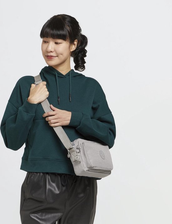 Een vrouw met een donkergroene hoodie en zwarte leren broek draagt een lichtgrijze Kipling schoudertas.