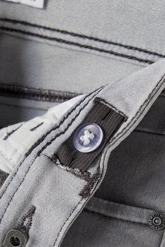 Detail van de tailleband van een grijze jeans, met verstelbare knoopsluiting en rits.