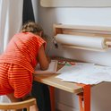 Kind met oranje gestreepte kleding zit aan tafel en tekent op roze lichttablet.