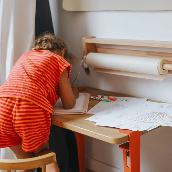 Kind met oranje gestreepte kleding zit aan tafel en tekent op roze lichttablet.