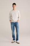 Man staand, dragend een beige trui en een blauwe jeans met lichte wassing.
