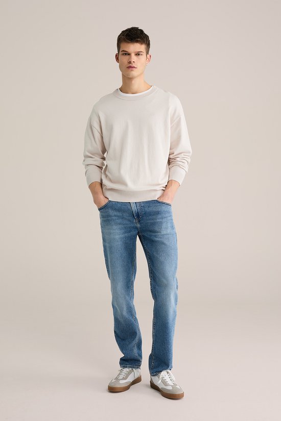Man staand, dragend een beige trui en een blauwe jeans met lichte wassing.