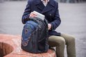 Man in donkerblauwe jas plaatst een laptop in een zwarte rugzak met blauwe en grijze vlakken.