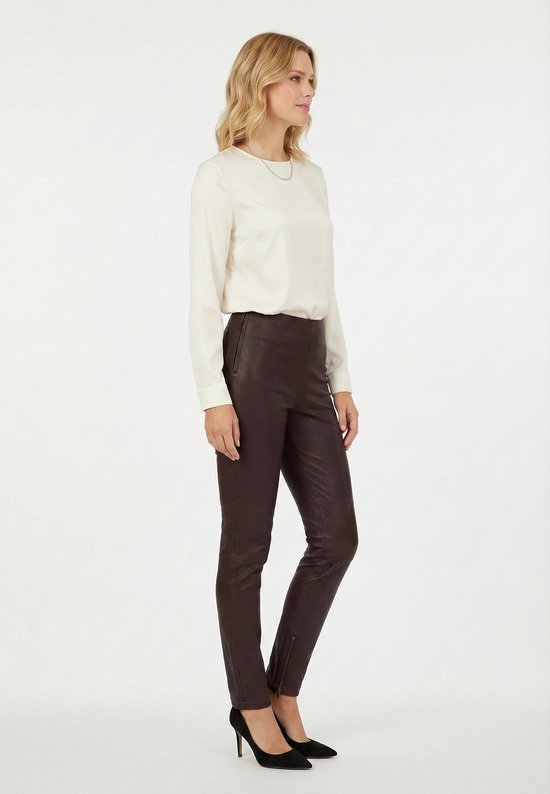 Vrouw met blond haar, profiel, draagt een beige blouse en een donkerbruine leren broek.
