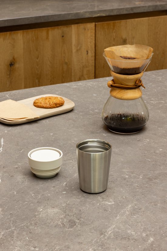 Zilverkleurige thermosbeker, los deksel en koffiezetapparaat met filter op een grijs aanrecht.