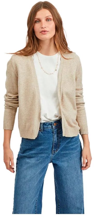 Een vrouw in een beige gebreid vest, wit T-shirt en spijkerbroek, staand.