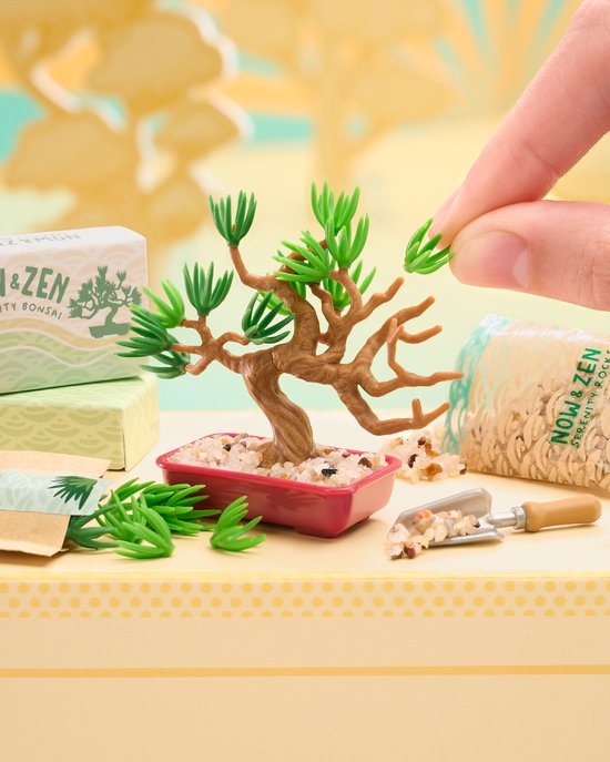 Vinger plaatst blaadjes op een miniatuur bonsaiboompje in een roze potje, met zakjes 'Now+Zen Serenity Rocks'.