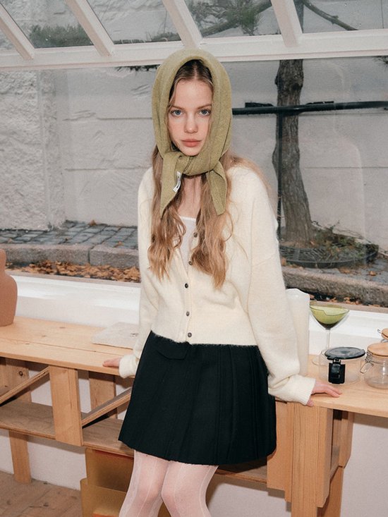 Model draagt een gebreide gebroken witte vest en een zwarte geplooide rok. De sjaal is in olijfgroen en wordt als hoofddoek gedragen.