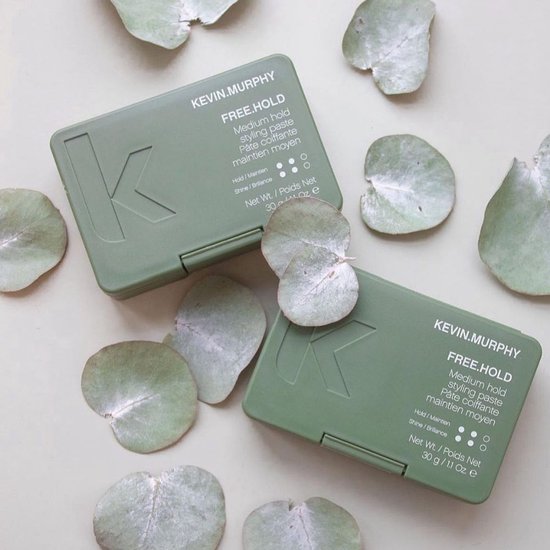 Twee groene KEVIN.MURPHY potjes met medium hold styling paste, omringd door eucalyptusblaadjes.