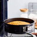 Zwarte ronde springvorm met goudkleurig gebakken cake, op een metalen rooster.
