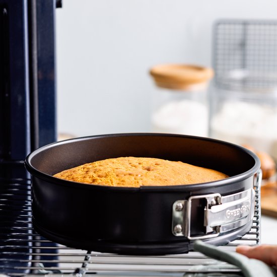 Zwarte ronde springvorm met goudkleurig gebakken cake, op een metalen rooster.