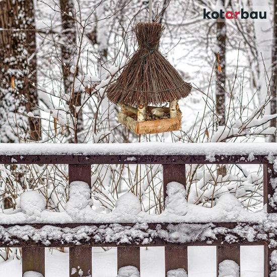 Houten en wilgen vogelhuisje hangend in de sneeuw, op een winterse achtergrond met houten hek.