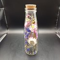 Glazen fles met kurk, gevuld met gedroogde bloemen in roze, paars, wit en blauw.