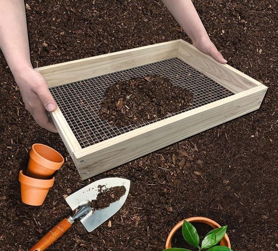 Een houten zeef met metalen gaas, gevuld met aarde. Twee kleine bloempotten en een schepje liggen ernaast.