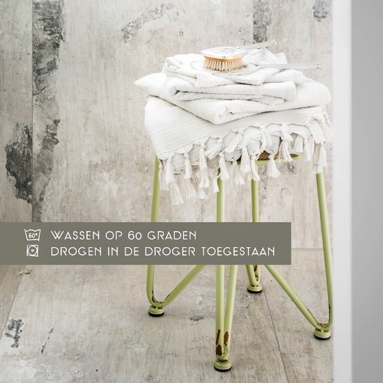 Stapel crèmekleurige handdoeken op een lichtgroene kruk. Tekst: WASSEN OP 60 GRADEN, DROGEN IN DE DROGER TOEGESTAAN.