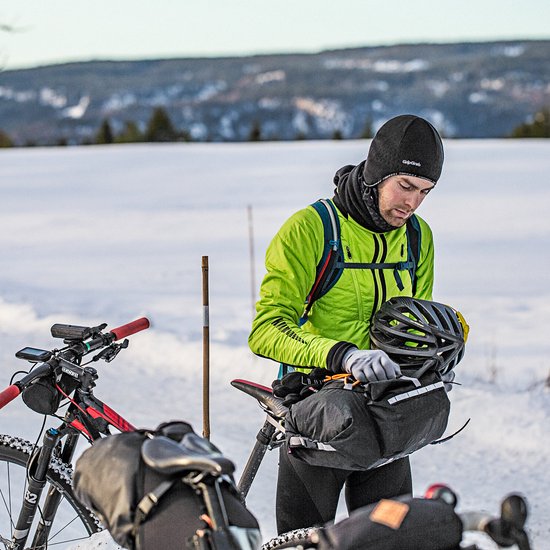 Man in een groen jack met zwarte fietsmuts en helm in besneeuwd landschap.