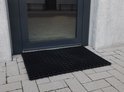 Zwarte ringmat met open structuur, bij de ingang van een gebouw.