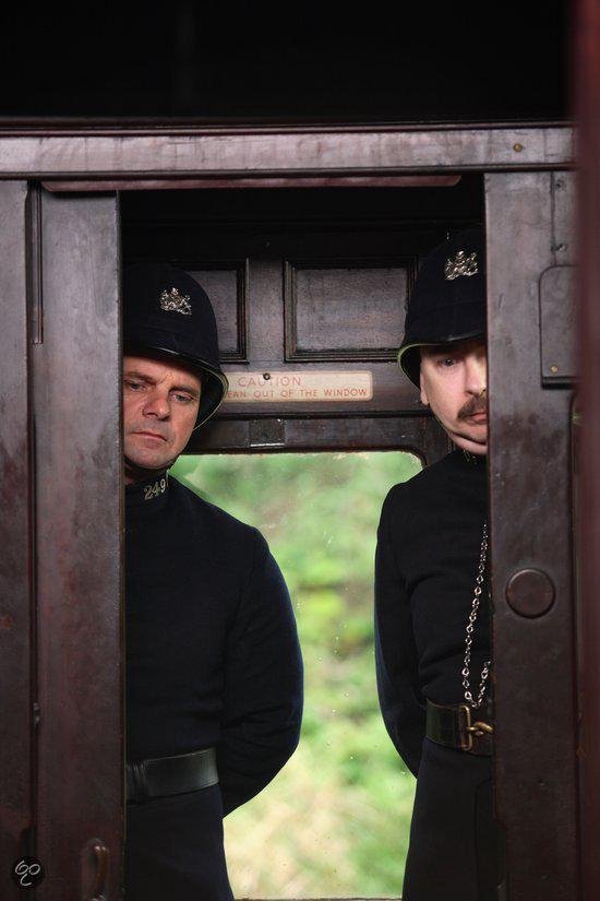Twee mannen in politieuniform met petten kijken vanuit een deuropening van een trein naar buiten.