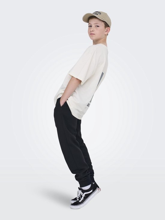 Jongen in crème T-shirt, zwarte joggingbroek en beige pet.