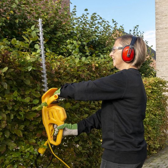 Vrouw met gehoorbescherming en veiligheidsbril snoeit een heg met een gele elektrische heggenschaar.