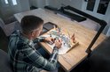 Man schildert miniaturen aan een houten tafel, verlicht door een zwarte bureaulamp.