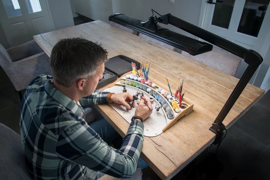 Man schildert miniaturen aan een houten tafel, verlicht door een zwarte bureaulamp.
