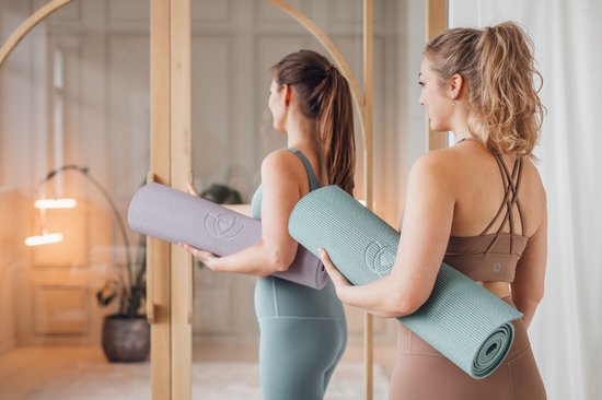 Twee vrouwen met sportkleding, de één draagt een paarse, de ander een kelmengroene yogamat.