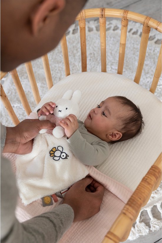 Een baby ligt in een wiegje met een gebreid wit knuffeldoekje met een vogeltje en een wit konijntje met een roze trui.