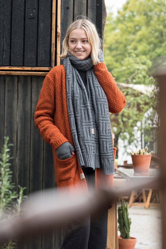 Model in oranje gebreide lange vest met grijze sjaal met patroon en bijpassende handschoenen.