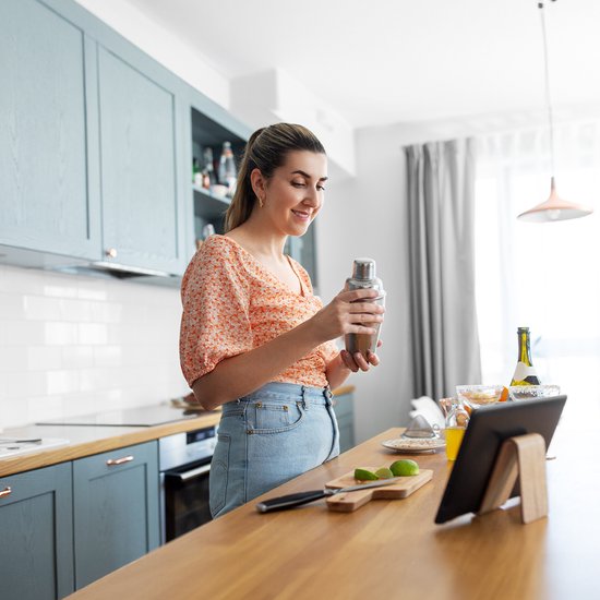 Een glimlachende vrouw met een oranje blouse en spijkerbroek houdt een cocktailshaker vast in een moderne keuken.